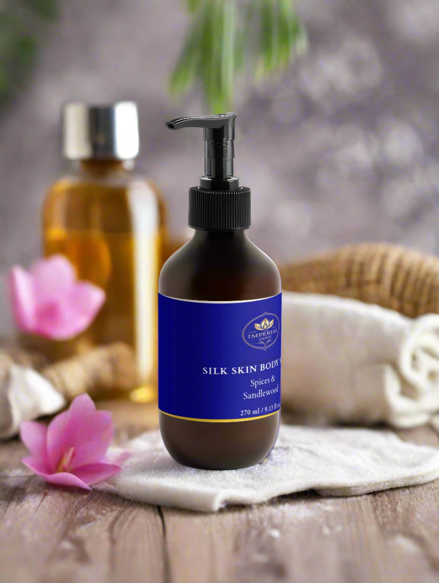 Bottle of Silk Skin Body product with a blue label on a wooden surface with flowers and another bottle in the background.